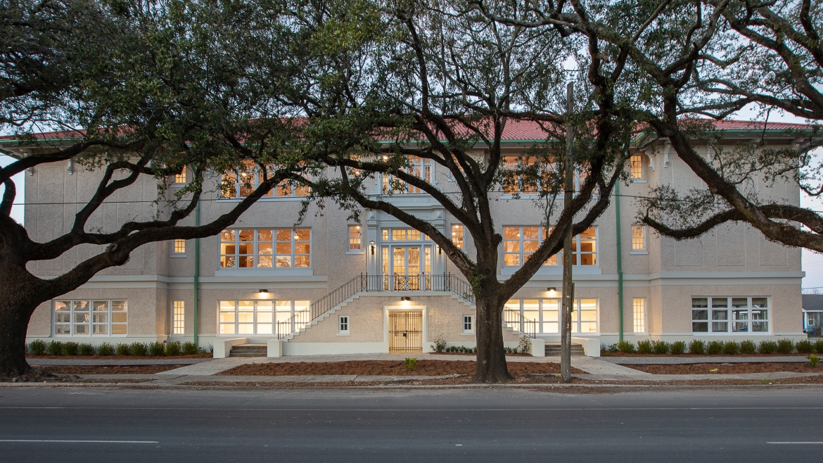 Exterior of a building at dusk with oak trees lining the sidewalk. 