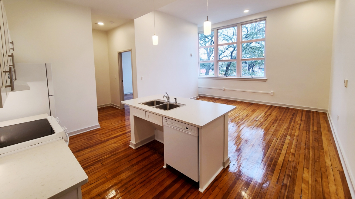 An empty apartment with wooden floors viewed from the kitchen looking into the living area.