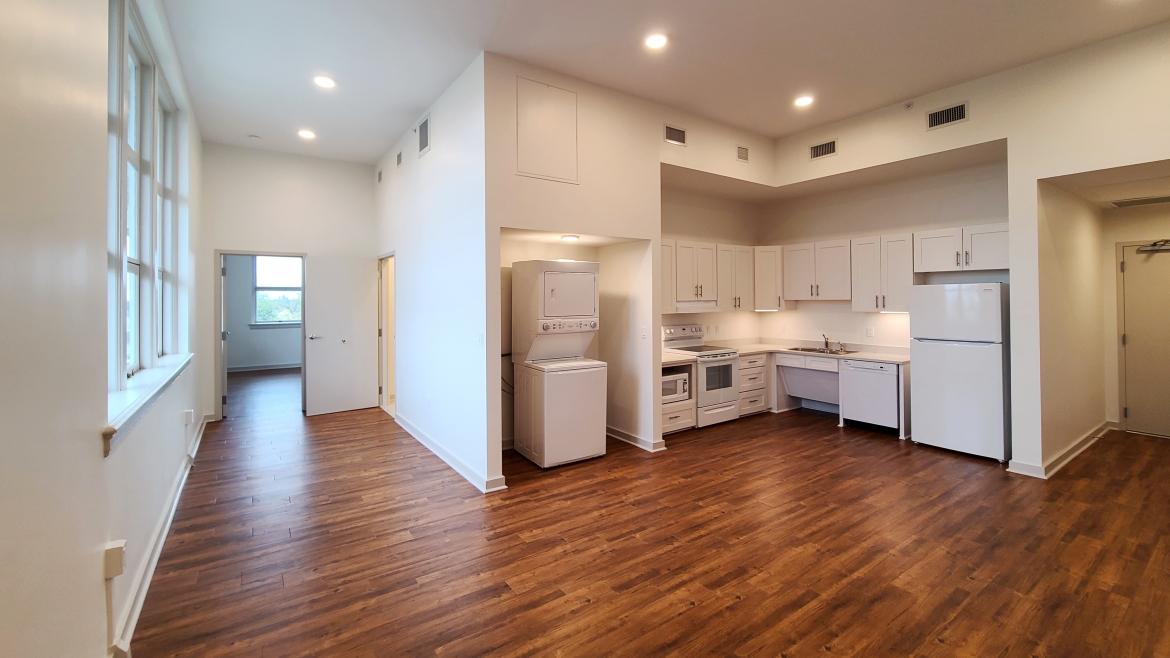 An empty apartment with wooden floors viewed from the living area looking toward the kitchen.